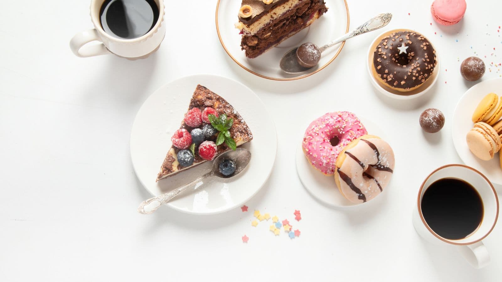 Assorted desserts including cake slices, donuts, macarons, chocolates, and two cups of black coffee, arranged on a white table with a few colorful star-shaped sprinkles scattered around.