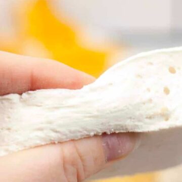 A hand holds a thick, white, soft rectangular piece of bread or dough, showing its airy texture and small holes. The background is blurred with yellow objects.