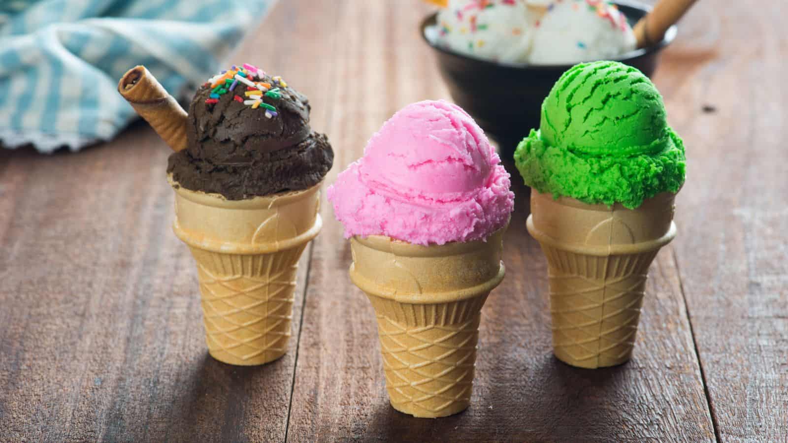 Three ice cream cones with chocolate, pink, and green scoops are arranged on a wooden table. The chocolate cone has sprinkles and a wafer stick. In the background, a bowl of ice cream scoops is partially visible.
