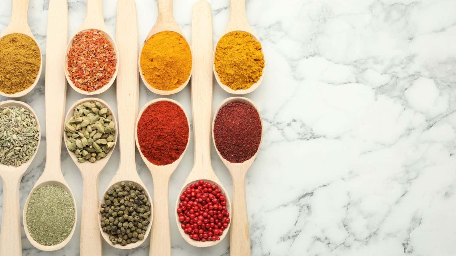 Wooden spoons filled with a variety of colorful spices, herbs, and peppercorns are arranged in neat rows on a white marble surface, with ample empty space on the right side.