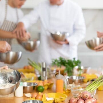 A group of people wearing striped aprons prepare food together in a kitchen, mixing ingredients in metal bowls. Various fresh vegetables, herbs, and kitchen tools are arranged on a wooden countertop.