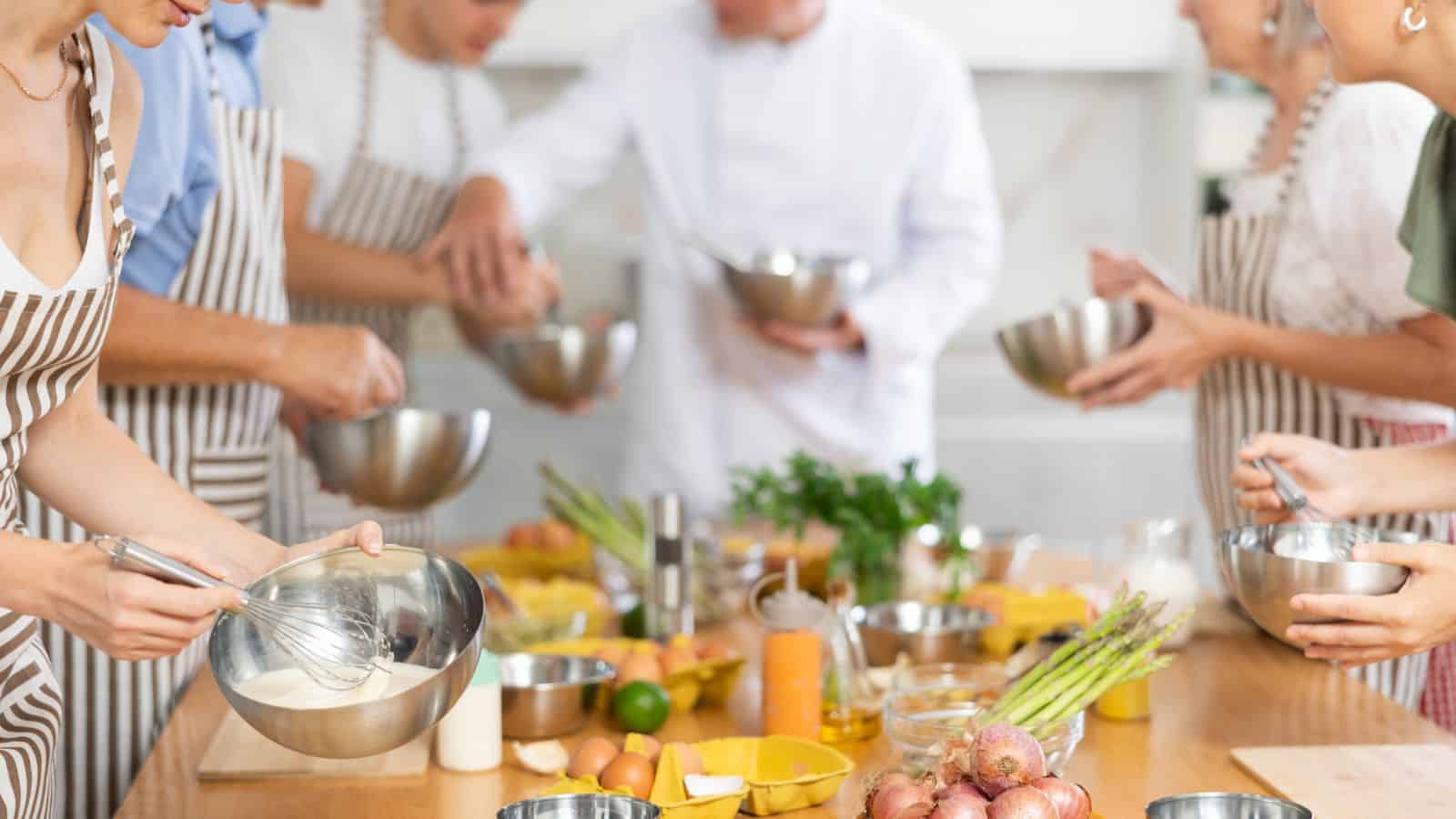 A group of people wearing striped aprons prepare food together in a kitchen, mixing ingredients in metal bowls. Various fresh vegetables, herbs, and kitchen tools are arranged on a wooden countertop.