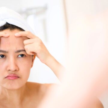 A person with a white towel wrapped around their head stands in front of a mirror, looking at their reflection and squeezing the skin on their forehead with both hands.