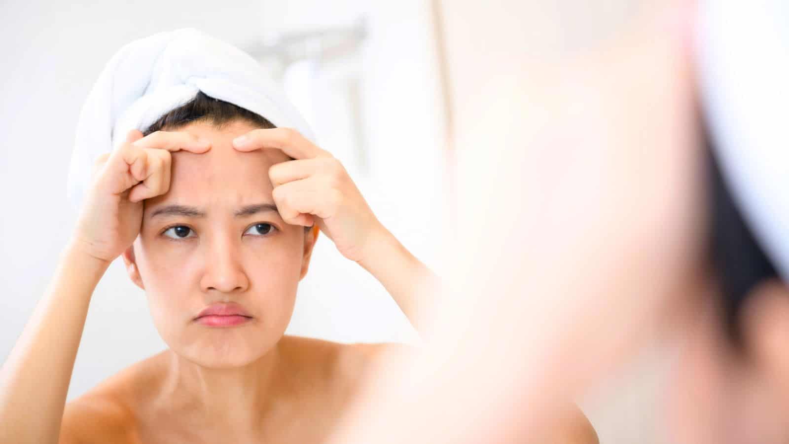 A person with a white towel wrapped around their head stands in front of a mirror, looking at their reflection and squeezing the skin on their forehead with both hands.