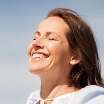 A woman with brown hair smiles with her eyes closed, facing toward the sunlight. She is wearing a light-colored top and appears to be outdoors with a blue sky in the background.