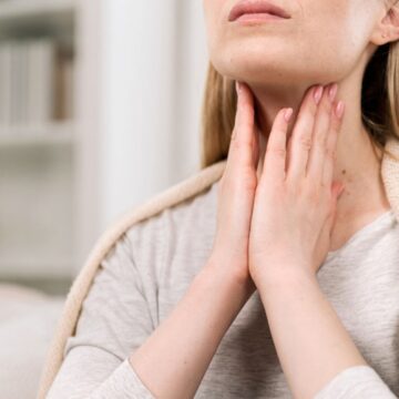 A person wearing a light sweater is sitting indoors and gently holding their throat with both hands, suggesting throat discomfort or pain. Shelves with books and decor are visible in the blurred background.
