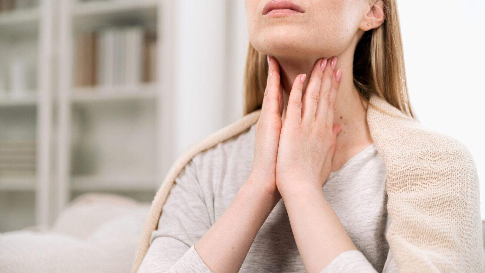 A person wearing a light sweater is sitting indoors and gently holding their throat with both hands, suggesting throat discomfort or pain. Shelves with books and decor are visible in the blurred background.