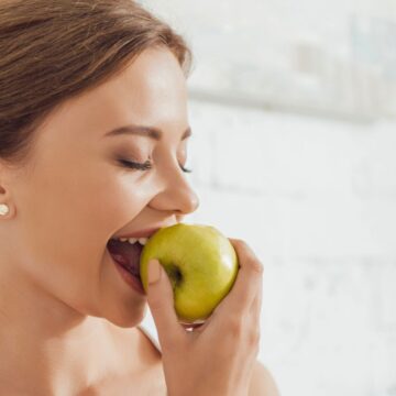 A woman with brown hair tied back is about to take a bite of a green apple. She is smiling with her eyes closed and standing in front of a white brick wall.
