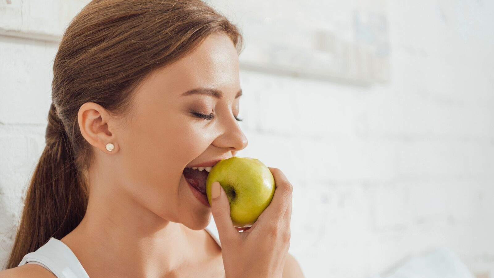 A woman with brown hair tied back is about to take a bite of a green apple. She is smiling with her eyes closed and standing in front of a white brick wall.
