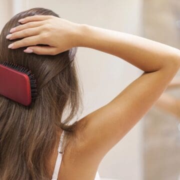 A woman stands in front of a mirror brushing her long brown hair with a red hairbrush, wearing a white tank top. Her arms are raised as she styles her hair in a bathroom setting.