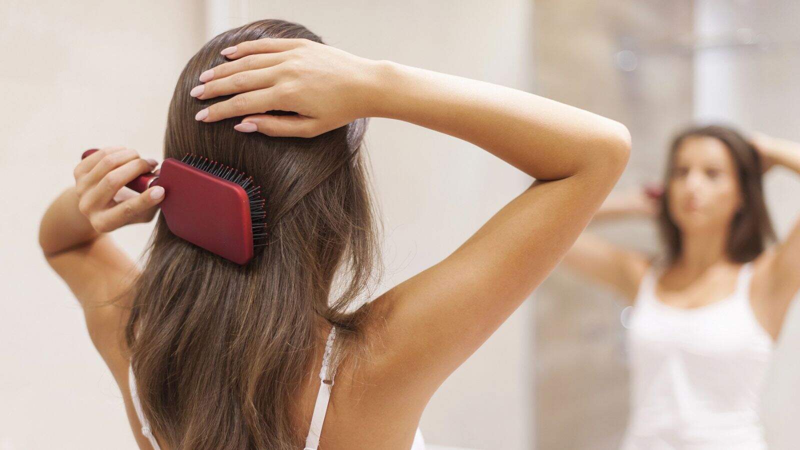 A woman stands in front of a mirror brushing her long brown hair with a red hairbrush, wearing a white tank top. Her arms are raised as she styles her hair in a bathroom setting.