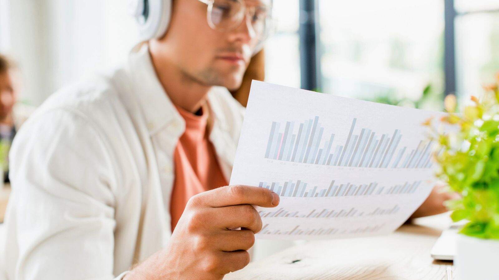 A person wearing headphones and glasses examines a printed sheet with bar graphs. The person is seated indoors near a bright window, with green plants visible in the foreground.