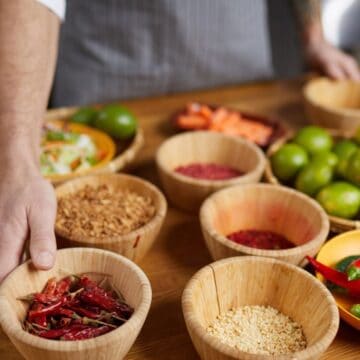 A person in an apron arranges wooden bowls containing dried red chilies, seeds, grains, fresh limes, and other ingredients on a wooden table. More food ingredients are visible in the background.