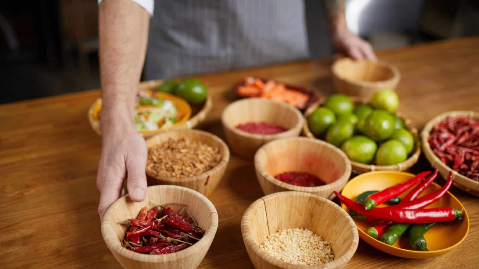 A person in an apron arranges wooden bowls containing dried red chilies, seeds, grains, fresh limes, and other ingredients on a wooden table. More food ingredients are visible in the background.