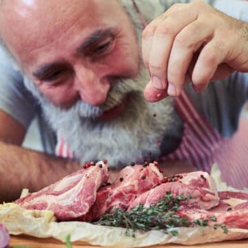 A man with a gray beard and striped apron sprinkles pepper onto raw lamb chops on parchment paper, surrounded by fresh vegetables such as bell pepper, broccoli, and carrots.