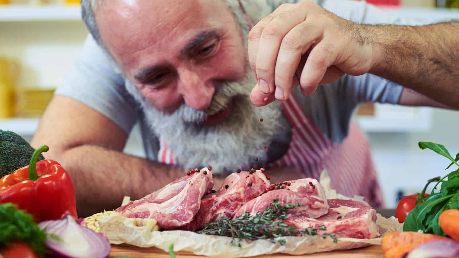A man with a gray beard and striped apron sprinkles pepper onto raw lamb chops on parchment paper, surrounded by fresh vegetables such as bell pepper, broccoli, and carrots.