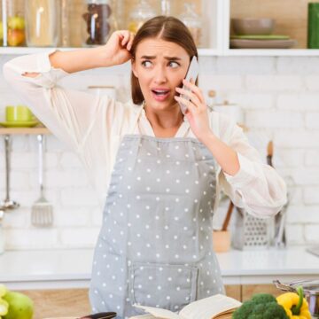 A woman wearing a gray polka dot apron talks on the phone in a kitchen, looking confused while scratching her head. An open cookbook, fruits, vegetables, and a pot are on the counter in front of her.