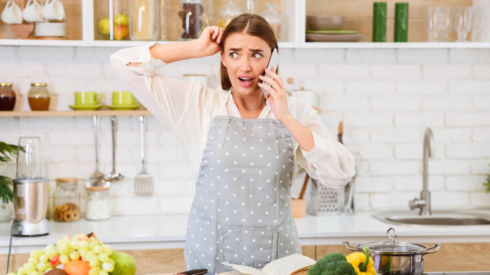 A woman wearing a gray polka dot apron talks on the phone in a kitchen, looking confused while scratching her head. An open cookbook, fruits, vegetables, and a pot are on the counter in front of her.