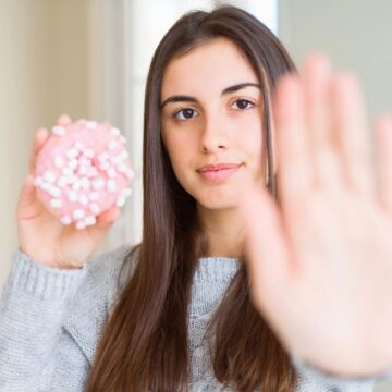A young woman with long brown hair holds a pink donut topped with mini marshmallows in one hand and raises her other hand in a stop gesture, facing the camera.