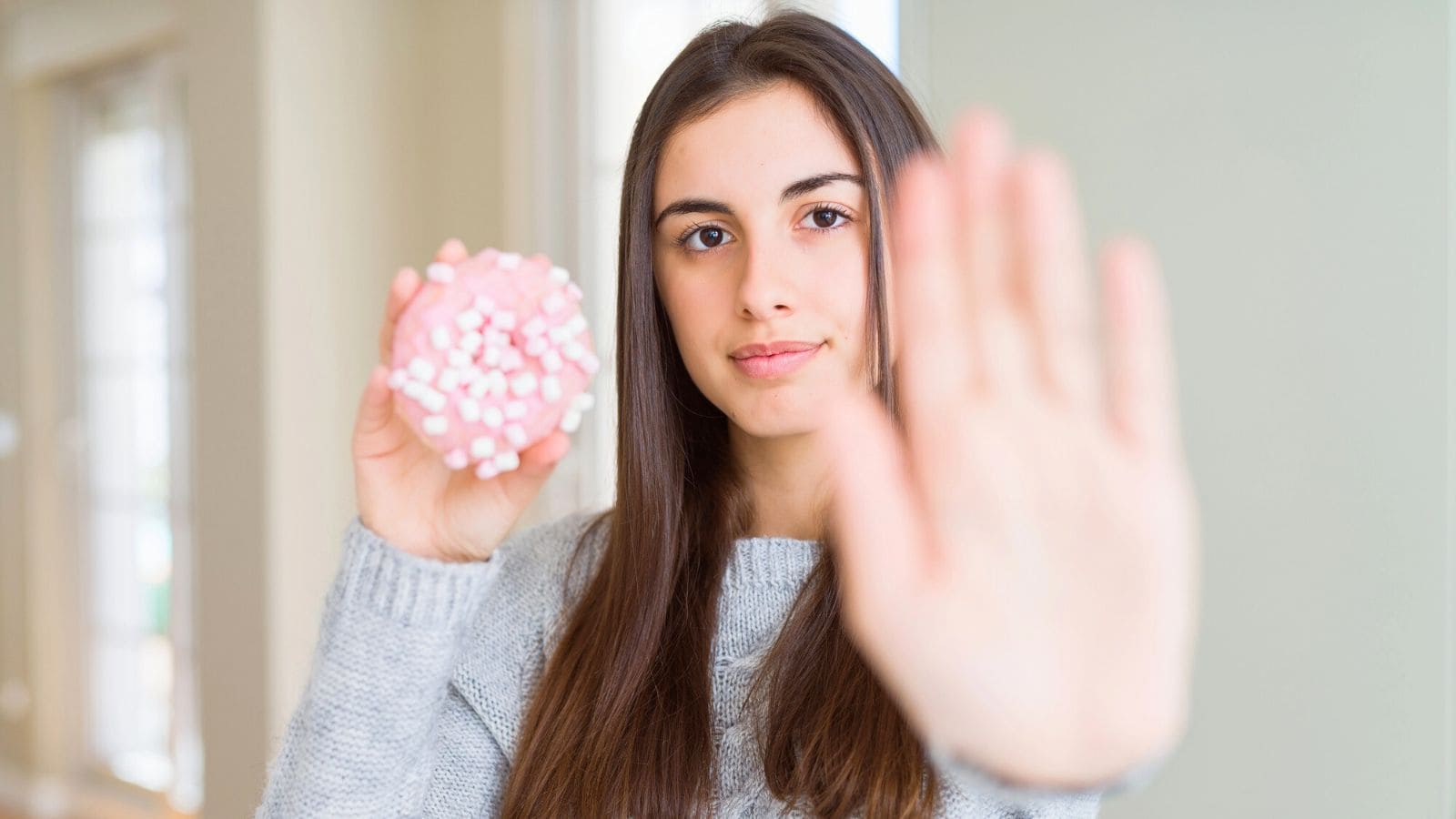 A young woman with long brown hair holds a pink donut topped with mini marshmallows in one hand and raises her other hand in a stop gesture, facing the camera.
