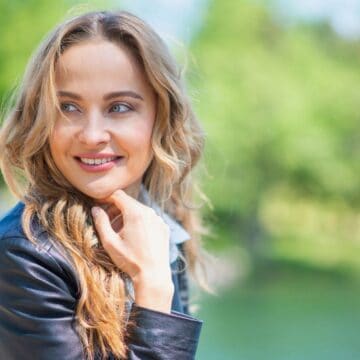 A woman with long, wavy blonde hair wearing a dark jacket smiles and looks to her left while standing outdoors near greenery and water on a sunny day.