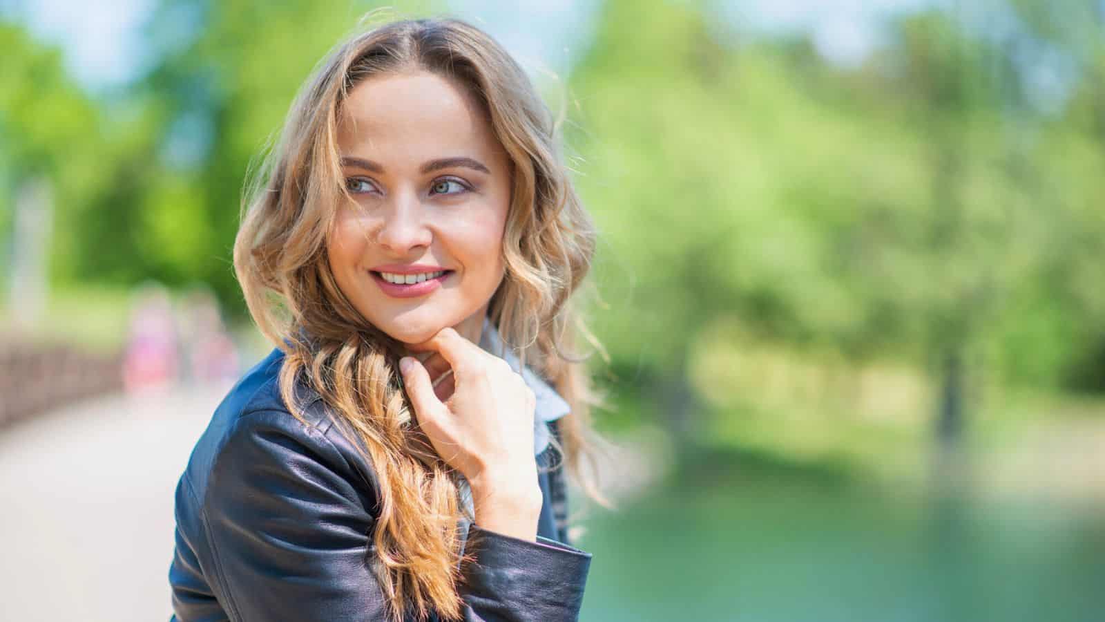 A woman with long, wavy blonde hair wearing a dark jacket smiles and looks to her left while standing outdoors near greenery and water on a sunny day.