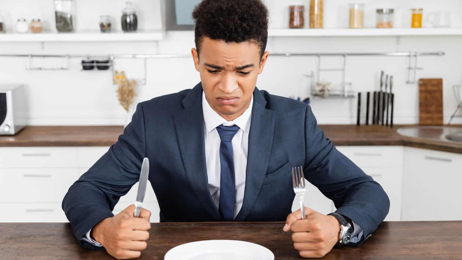 A man in a suit sits at a kitchen table holding a knife and fork, looking disappointedly at an empty white plate in front of him. The kitchen background includes shelves with jars and utensils.
