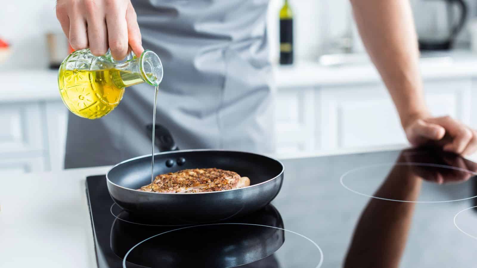 A person wearing a grey apron pours olive oil from a glass pitcher into a frying pan with a seasoned piece of meat on an electric stovetop in a bright kitchen.