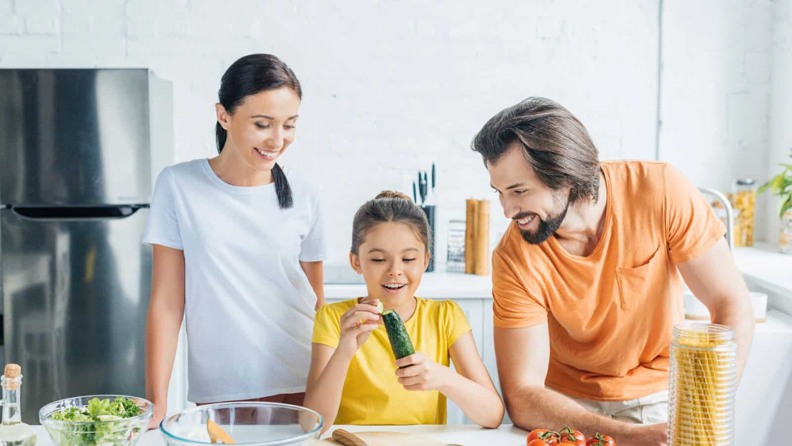 A woman, a man, and a young girl are standing in a kitchen. The girl is holding a cucumber and smiling, while the adults watch her. Various vegetables and kitchen items are on the counter in front of them.