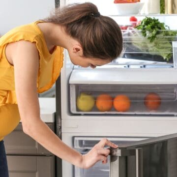 A woman in a yellow top stands in front of an open refrigerator, looking inside. The fridge contains fresh produce, bottles, and jars, with fruits visible in the bottom drawers. She is opening the lower freezer compartment.