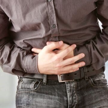 A person wearing a gray button-up shirt and jeans is holding their stomach with both hands, suggesting discomfort or pain in the abdominal area.