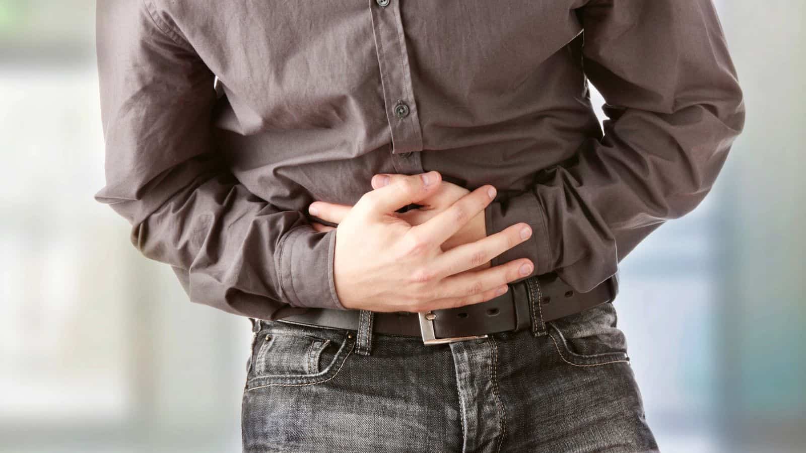 A person wearing a gray button-up shirt and jeans is holding their stomach with both hands, suggesting discomfort or pain in the abdominal area.