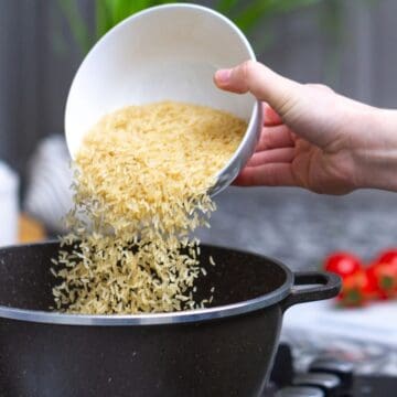 A hand is pouring uncooked rice from a white bowl into a black pot on a stovetop in a kitchen. Some fresh tomatoes and herbs are visible in the blurred background.