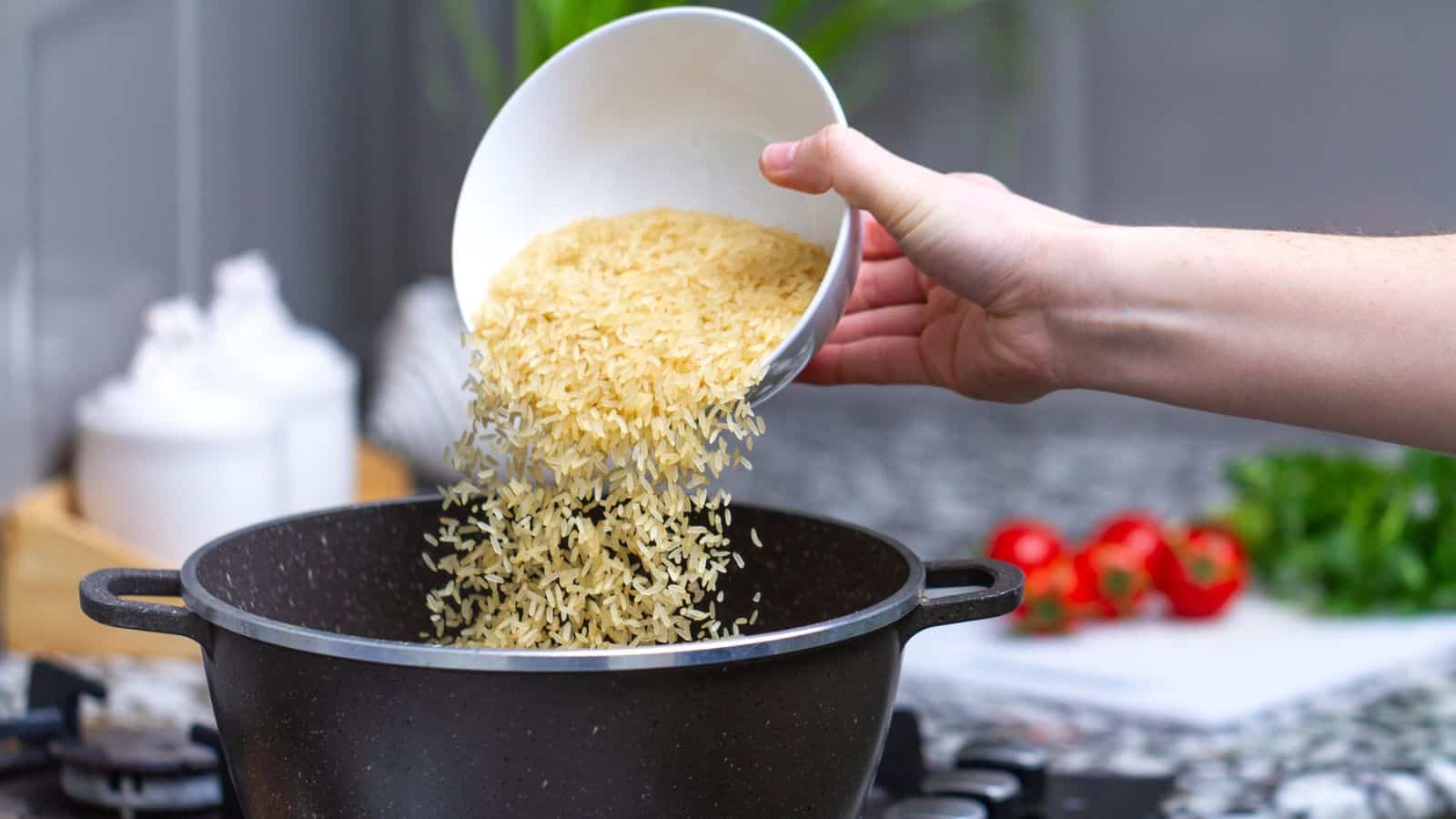 A hand is pouring uncooked rice from a white bowl into a black pot on a stovetop in a kitchen. Some fresh tomatoes and herbs are visible in the blurred background.