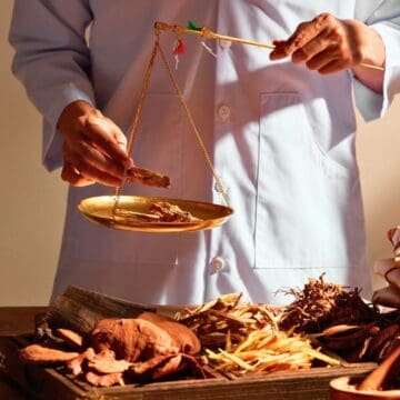 A person in a light blue coat uses a balance scale to weigh dried herbs on a table covered with various traditional medicinal ingredients, a mortar and pestle, and tied packages.