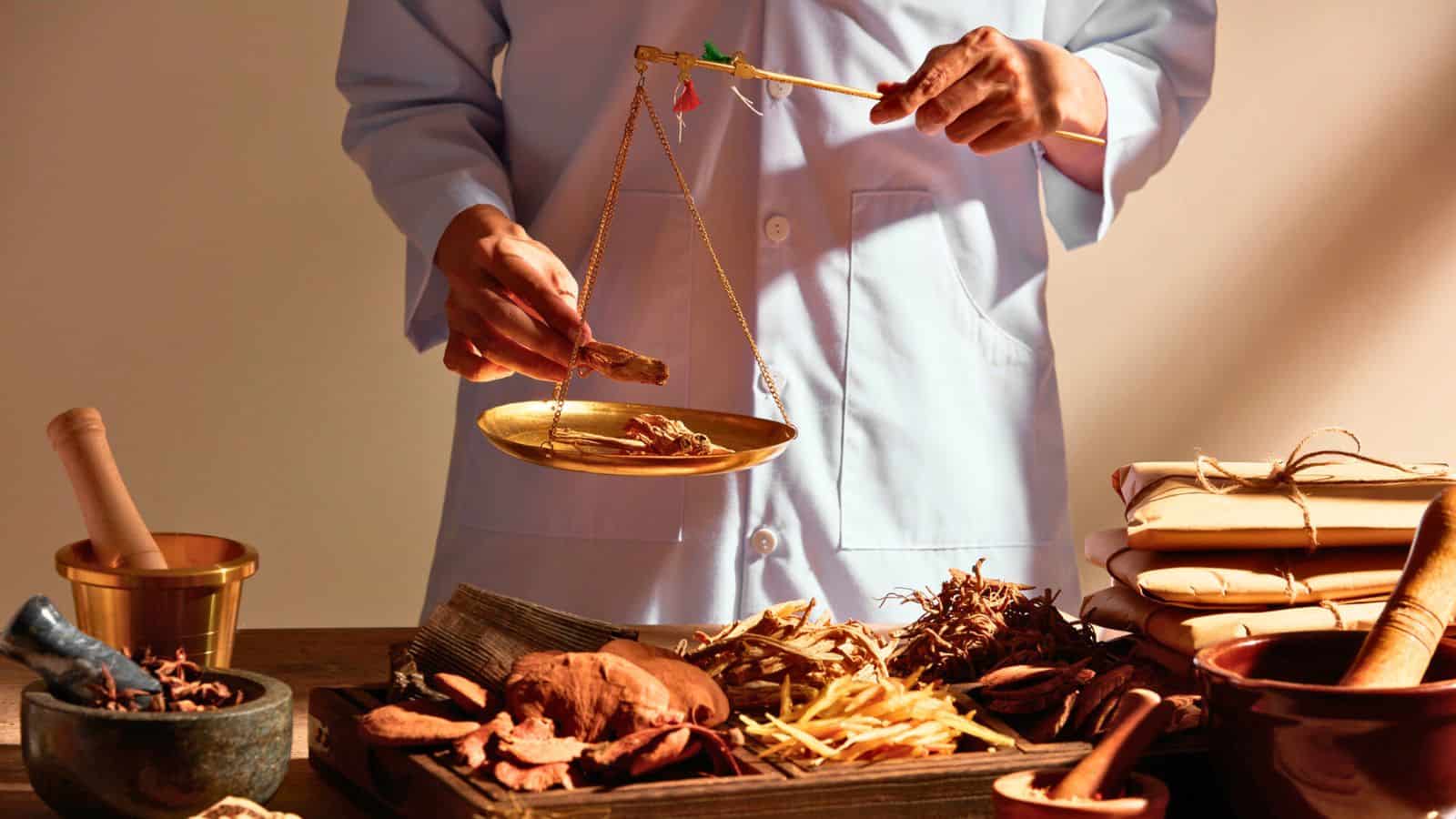 A person in a light blue coat uses a balance scale to weigh dried herbs on a table covered with various traditional medicinal ingredients, a mortar and pestle, and tied packages.