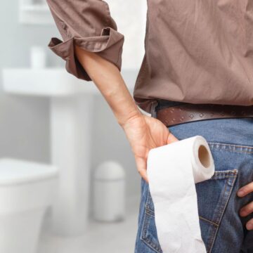 A person standing in a bathroom with their back to the camera, holding a roll of toilet paper in one hand and touching their backside with the other hand. A toilet and sink are visible in the background.