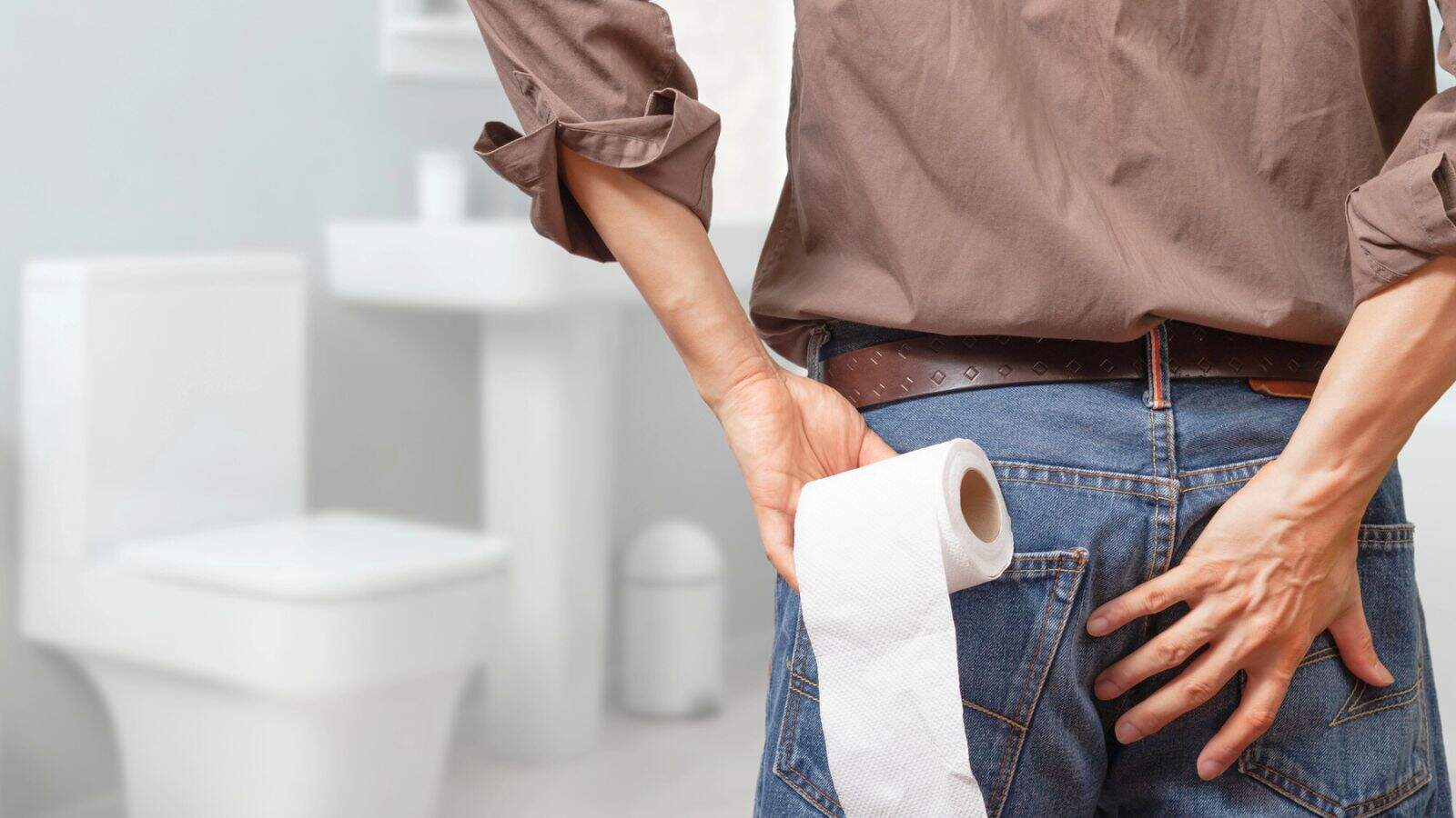 A person standing in a bathroom with their back to the camera, holding a roll of toilet paper in one hand and touching their backside with the other hand. A toilet and sink are visible in the background.