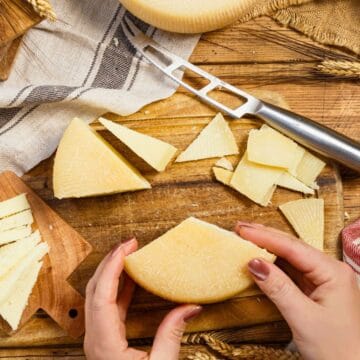 A person holds a round cheese wheel on a wooden table with sliced cheese, a cheese knife, cutting boards, wheat stalks, and cloth napkins arranged around it.