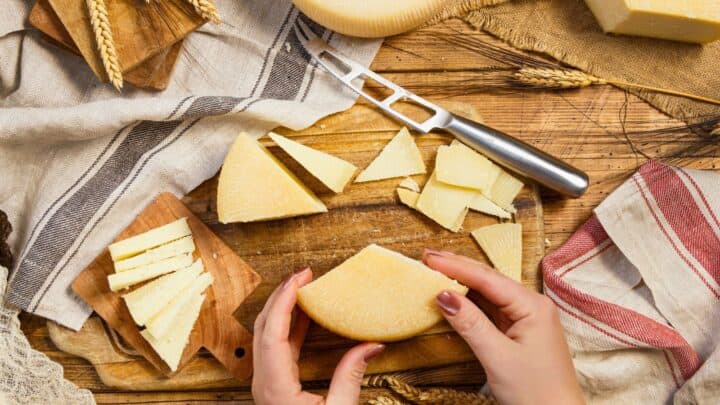 A person holds a round cheese wheel on a wooden table with sliced cheese, a cheese knife, cutting boards, wheat stalks, and cloth napkins arranged around it.