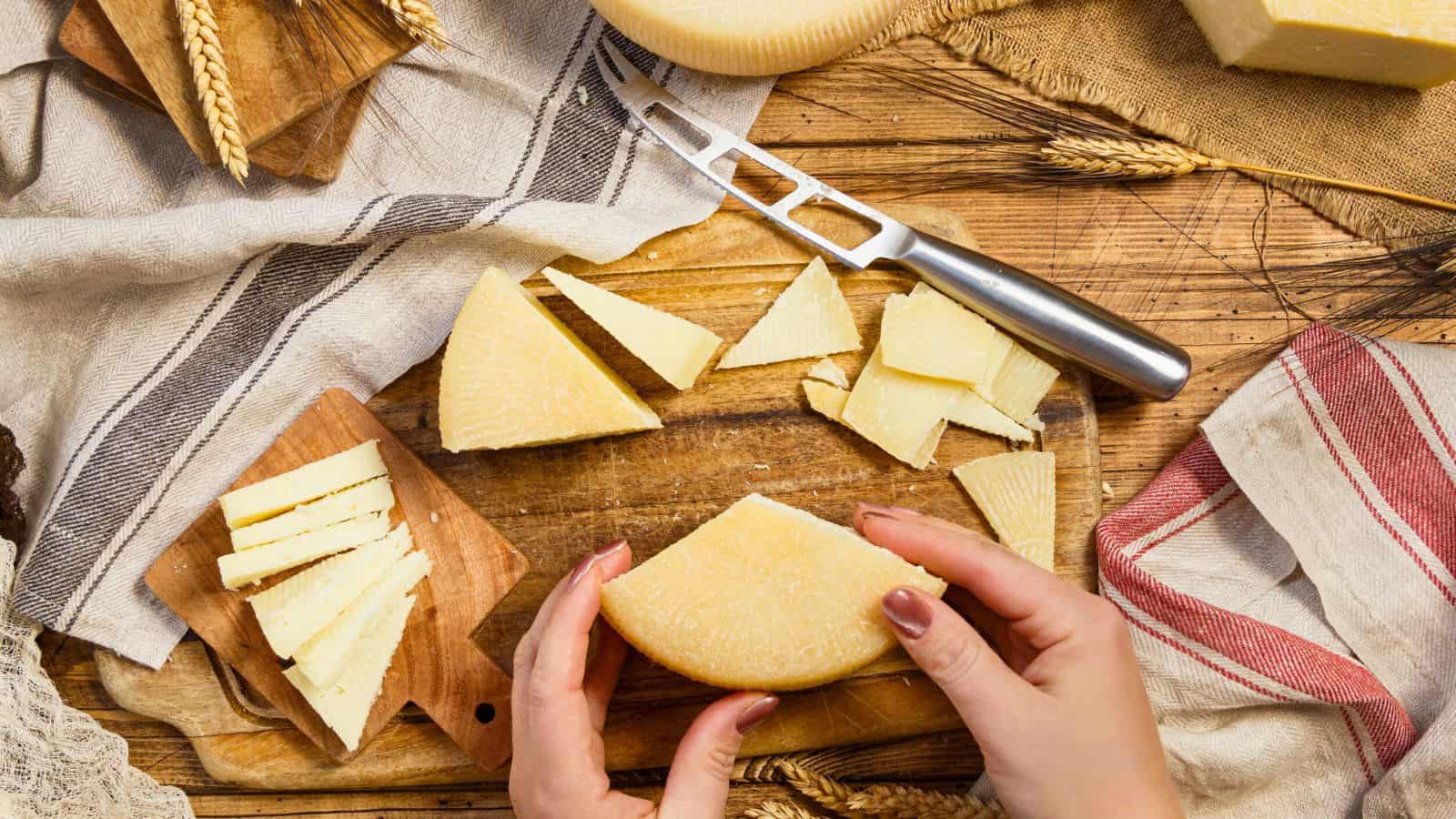 A person holds a round cheese wheel on a wooden table with sliced cheese, a cheese knife, cutting boards, wheat stalks, and cloth napkins arranged around it.