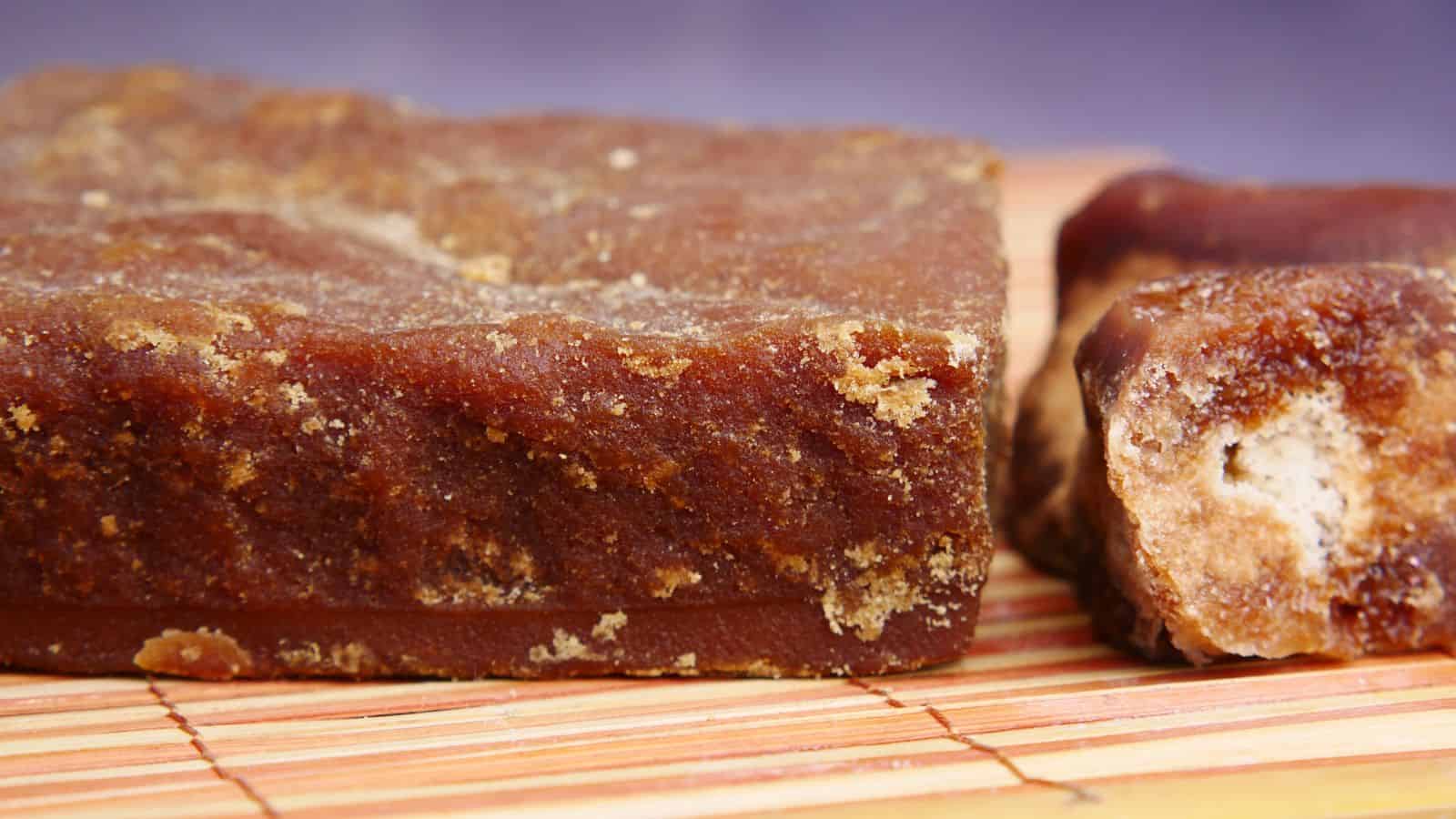 A close-up view of a block of brown jaggery with a rough texture, placed on a bamboo mat, next to a smaller broken piece showing its granular interior.
