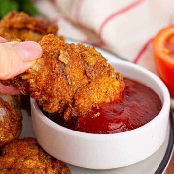 A hand dips a piece of fried chicken into a small bowl of red sauce, with more pieces of fried chicken and a halved tomato visible nearby.