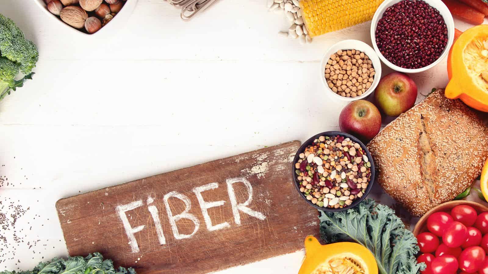 A wooden board with the word "FIBER" written on it is surrounded by high-fiber foods, including grains, beans, apples, broccoli, nuts, leafy greens, bread, corn, and squash, all arranged on a white surface.