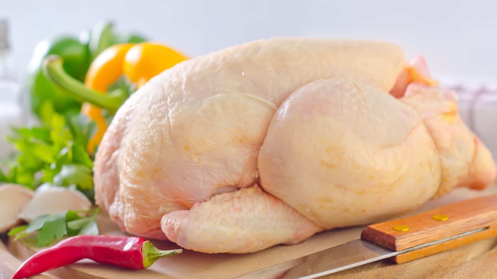 A raw whole chicken is placed on a wooden cutting board. Fresh vegetables, including parsley, a green pepper, a yellow bell pepper, garlic cloves, and a red chili pepper, are arranged around it. A knife is nearby.