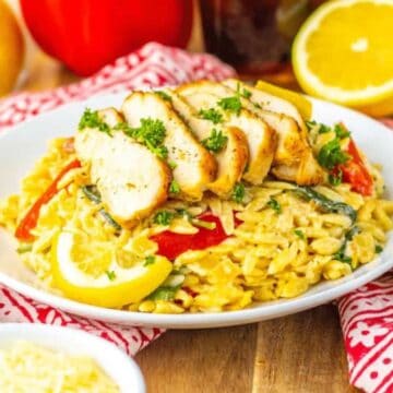 A plate of orzo pasta with creamy sauce, sliced grilled chicken, red bell pepper, spinach, and lemon wedges, garnished with parsley. Lemons, a red pepper, and parmesan cheese are in the background.