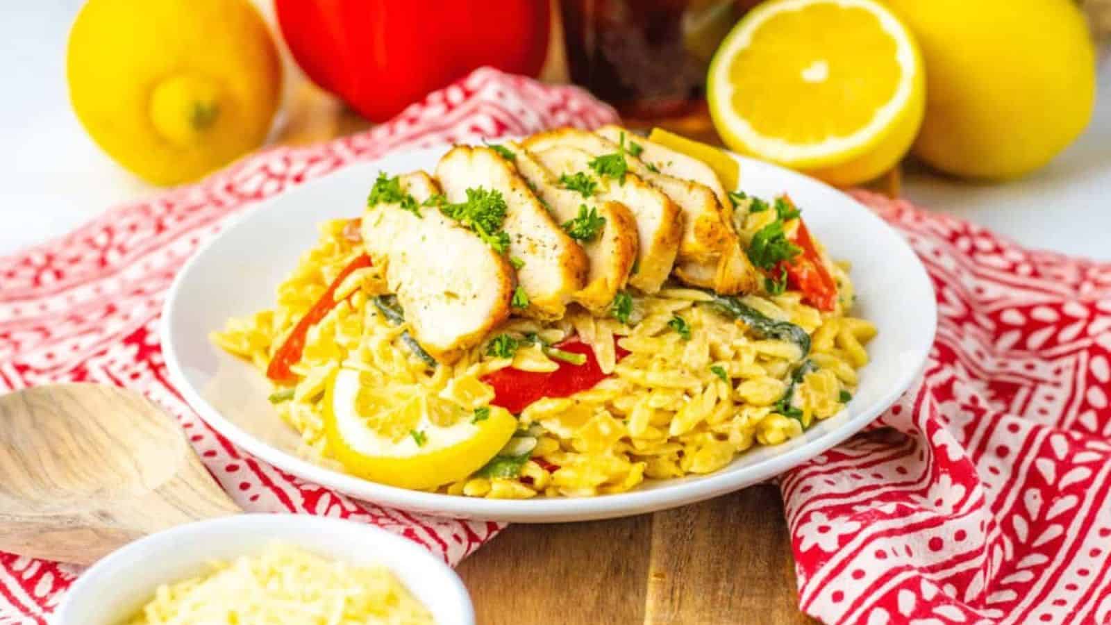 A plate of orzo pasta topped with sliced grilled chicken, garnished with parsley and lemon. Nearby are bell peppers, lemons, and a bowl of grated cheese on a red patterned cloth.