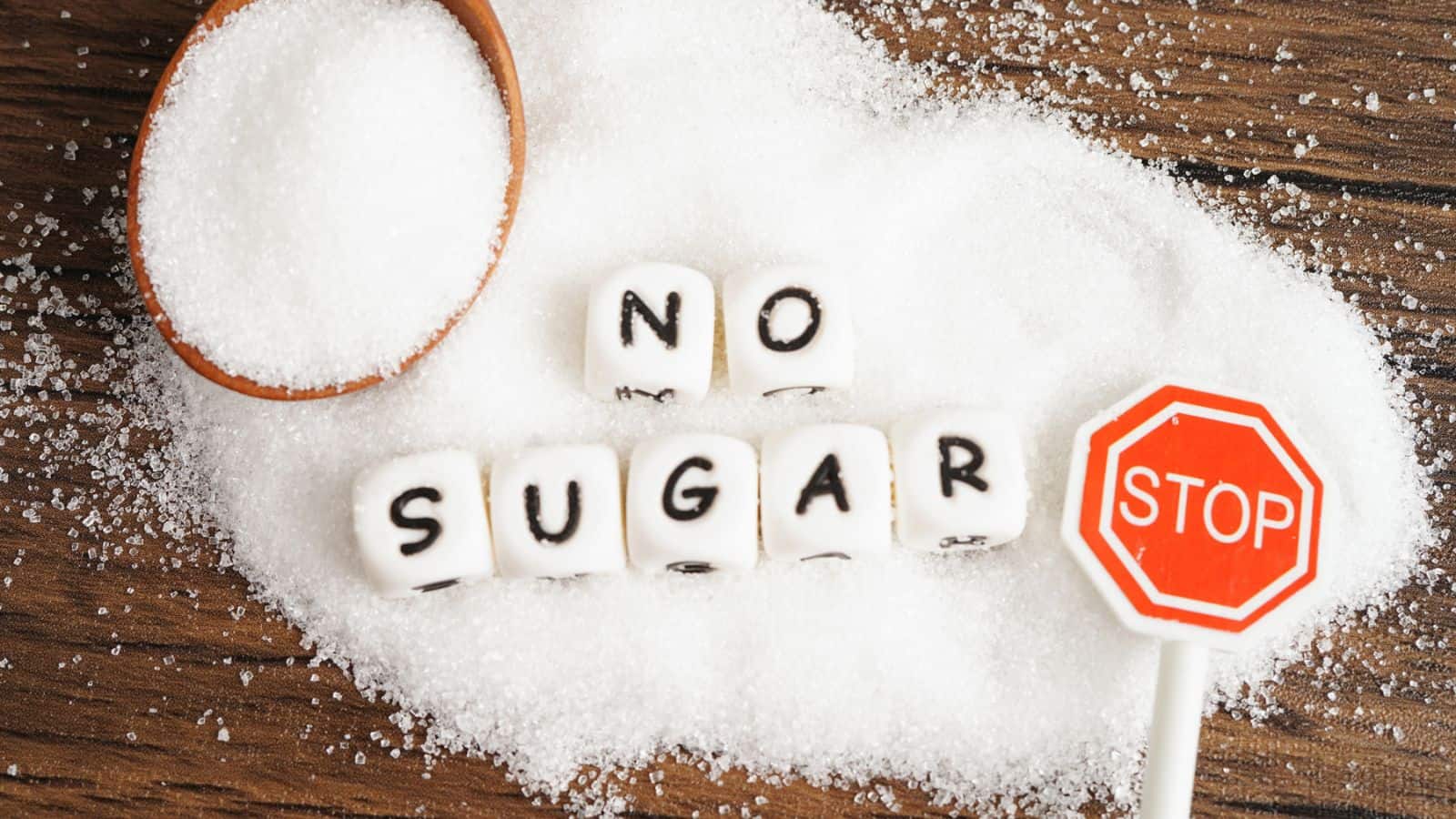 A wooden spoon with granulated sugar, sugar cubes spelling "NO SUGAR," and a small stop sign lollipop are arranged on a mound of sugar on a wooden surface.