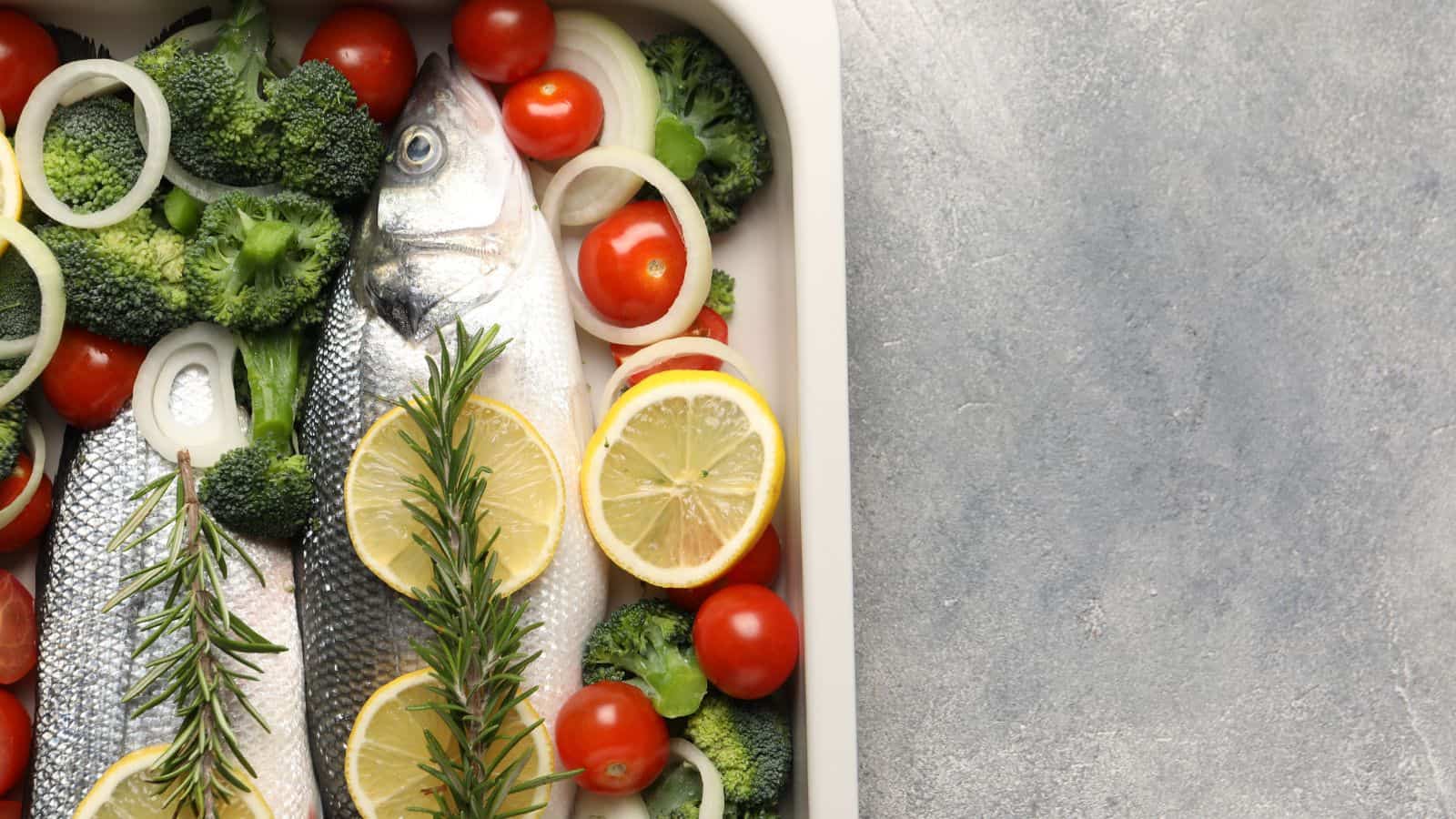 Two whole raw fish in a baking dish with lemon slices, rosemary, broccoli, cherry tomatoes, and onion rings, set against a gray textured surface.