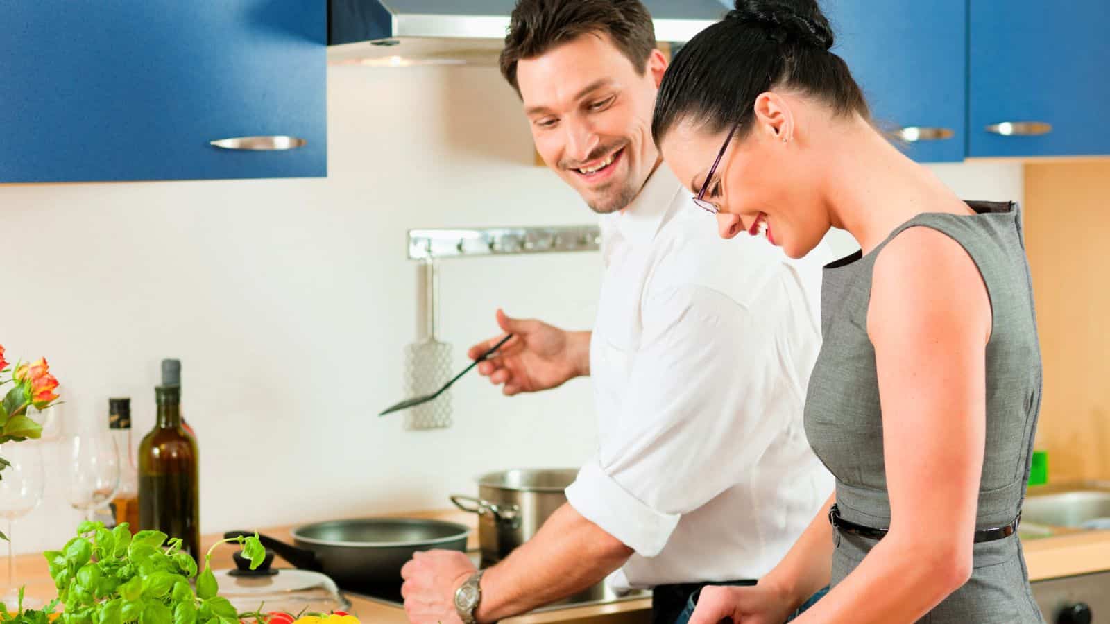 A man and a woman stand in a modern kitchen cooking together. The man holds a spatula by the stove, while the woman prepares food at the counter nearby. Various kitchen items and ingredients are visible.